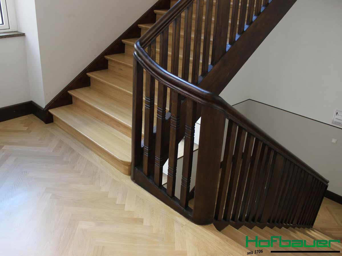 Weitere Ansicht des Treppengeländer aus dunklem Holz in der Nahaufnahme. Die Treppen sind in einem hellen Holz und einem Treppenabsatz in Fischgrätenoptik gestaltet
