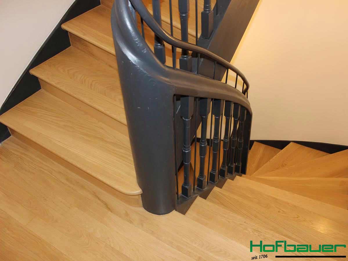 Seitliche Ansicht: Wendeltreppe aus hellem Massivholz mit einem schwarzen, ebenfalls aus Holz gefertigten Treppengeländer